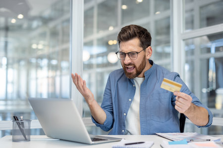 A frustrated businessman in a modern office, holding a credit card and staring at his laptop with an unhappy expression.の写真素材