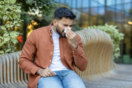 A man with a beard sits on a wooden bench outside, blowing his nose into a tissue during the day. The background features greenery.の写真素材