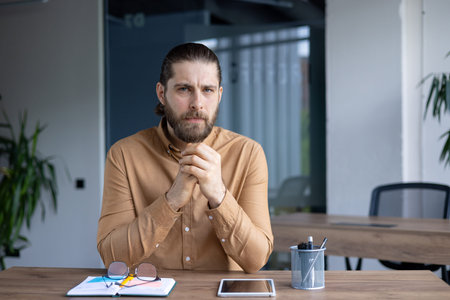A thoughtful man with a beard, looking directly at the camera. He is sitting at a wooden desk with a...