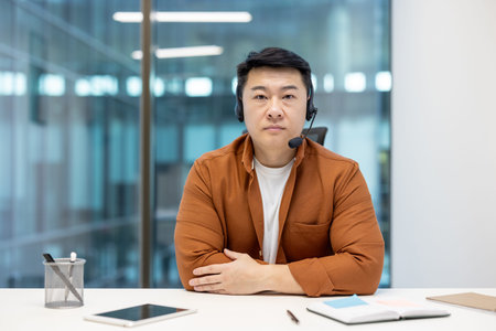 An asian businessman wearing a headset, sitting at his desk, looks directly at the camera with a...