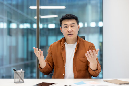 An asian businessman gestures while talking, captured in a bright office setting. He is wearing a casual orange shirt.の写真素材