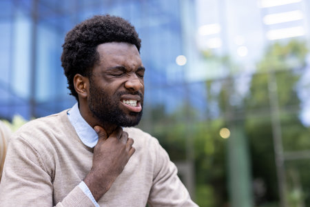 An african american man holds his neck, looking pained, suggesting a sore throat or other medical...