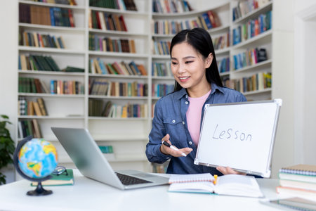 An asian woman gives an online lesson while holding a whiteboard with the word lesson in front of a laptop.の写真素材