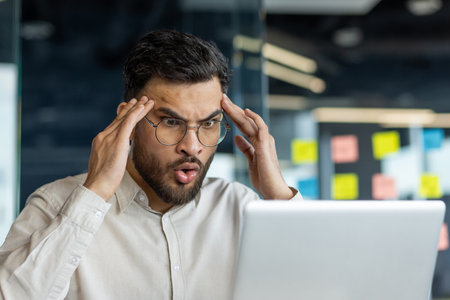 A shocked man stares at his laptop, showing an expression of surprise and concern while touching his temples. Office setting is visible in the background.の写真素材