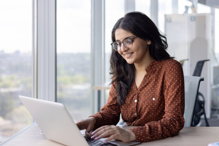 A smiling woman with glasses works on a laptop in a modern office setting. The office has a large window with a city view in the background.の写真素材