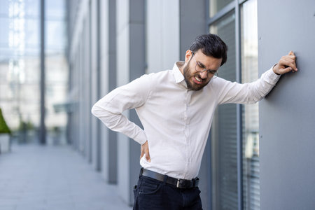 A businessman in a white shirt, glasses, and a beard is experiencing lower back pain outdoors near a...