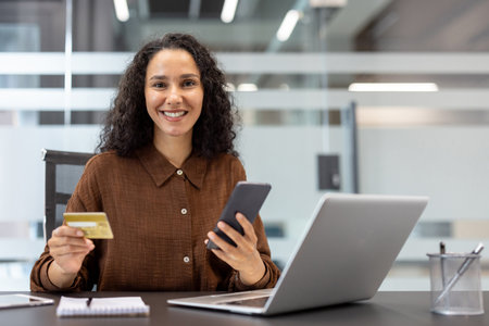 A smiling woman in a brown shirt holds a credit card and smartphone while seated at her desk with a laptop.の写真素材