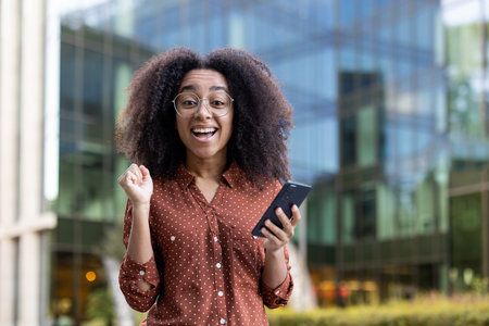 Excited young woman looks at her phone and celebrates a good news. A happy lady with glasses outside in front of a modern building.の写真素材