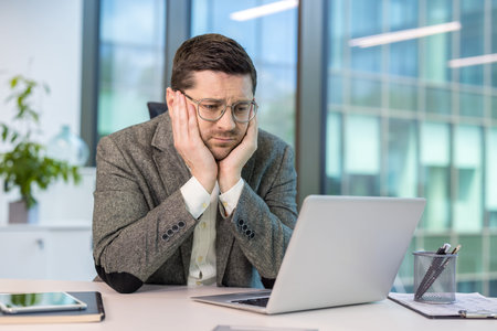 A tired and depressed young male office worker in a suit sits at his desk and looks boredly at the...