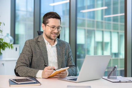 A smiling businessman works on a laptop in a modern office, writing in a notepad with a pen. Modern...
