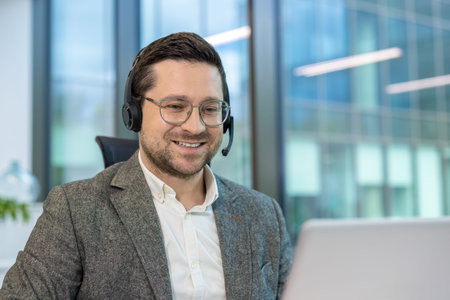 A close-up photo of a young man in a suit and headset sitting in an office at a desk ,and working on...