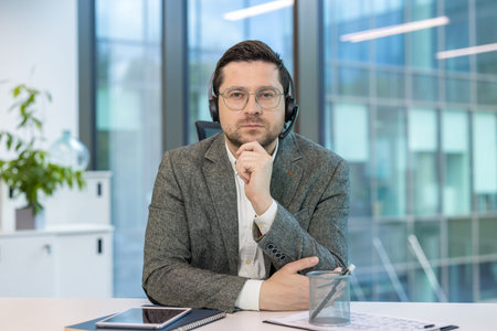 Portrait of a serious young man in a suit and headset sitting in the office at his desk, looking...