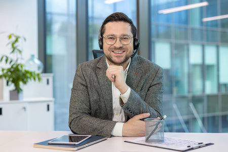 Portrait of a smiling young businessman in a headset and suit sitting at a desk in the office,...