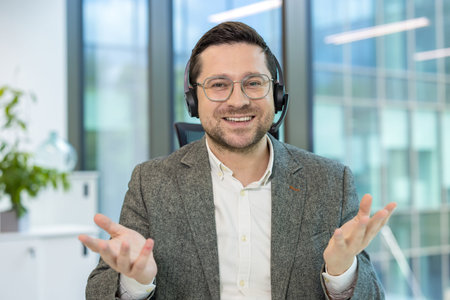 Portrait of a smiling young man in a headset sitting at an office desk and talking to the camera,...