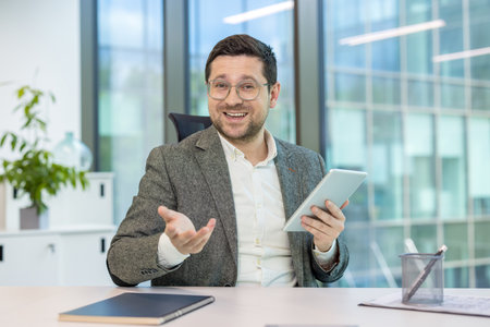 A businessman in a video conference, holding a tablet and gesturing in an office setting. He appears to be in a virtual meetingの写真素材