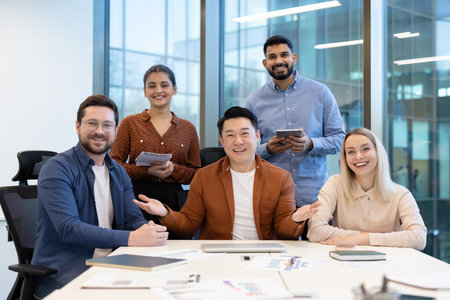 A diverse team smiles at the camera while gathered around a table in a modern office setting. Collaboration and teamwork are evident.の写真素材
