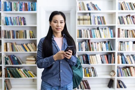 An asian woman is smiling while holding her phone in a library with many books in the background.の写真素材