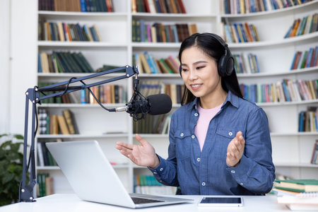 An asian woman records a podcast in front of a bookcase. She wears headphones and talks into a...