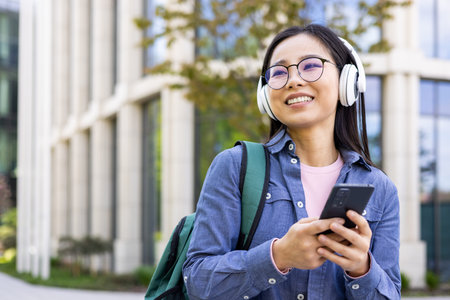 An asian student, wearing glasses and headphones, smiles while looking at her phone outdoors. She is...