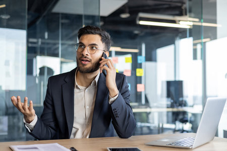 Serious confident businessman talking on the phone while sitting at the desk in the office....