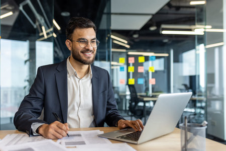A smiling businessman in glasses works on a laptop and documents in a modern office setting,...