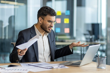 A frustrated businessman is looking at his laptop screen in the office, holding papers and gesturing...
