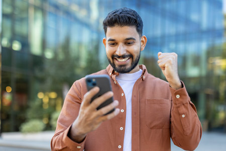 A young Indian man smiles with excitement while looking at his phone, celebrating success in front...