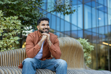 A cheerful man sits on a modern bench in front of a building, smiling and looking to the side.
