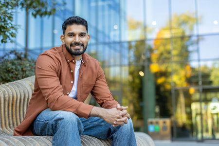 A smiling man is sitting on a bench with a modern building in the background, looking directly at...