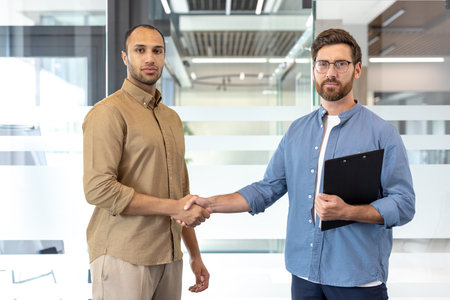 Two businessmen shake hands in an office setting, symbolizing a deal or agreement. The men are well-dressed and appear confident, set against a modern office environment.の写真素材