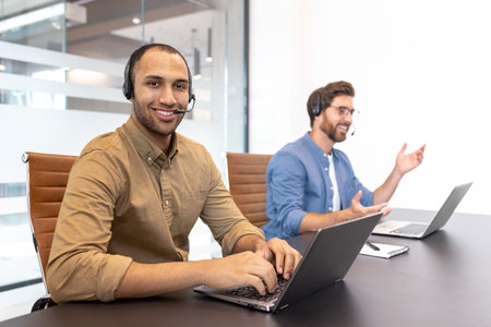 Two diverse customer service representatives wearing headsets are working on laptops in a modern...