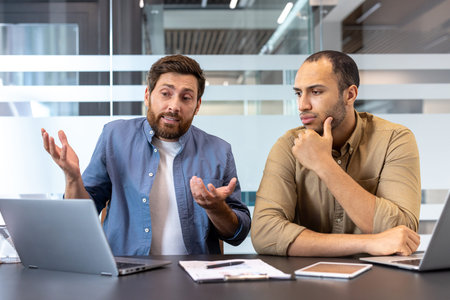 Two diverse business professionals discuss something in a modern office environment, using a laptop...