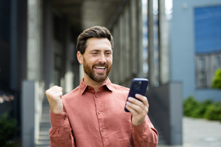 A man in a red shirt beams with joy as he checks his phone, clenching his fist in excitement outdoors.の写真素材