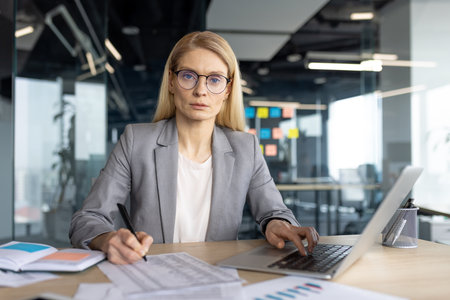 A professional businesswoman in a modern office setting, working on a laptop and taking notes, with a serious expression.の写真素材
