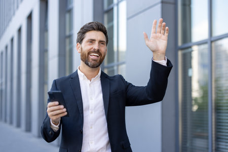 A businessman is waving while holding his phone in an urban setting. He looks happy and successful in his professional attire.の写真素材