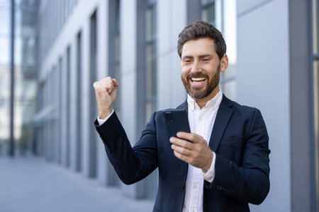 A successful businessman celebrates a win, looking at his phone, with a raised fist and a joyful expression near a modern building.の写真素材