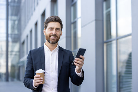 A smiling businessman in a suit stands outside a modern building, holding a coffee cup and smartphone.の写真素材