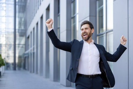 A businessman celebrates a victory, expressing joy with raised arms and an open-mouthed expression, outdoors.の写真素材