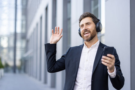 A smiling businessman in a suit waves while listening to music on headphones outside a modern building.の写真素材