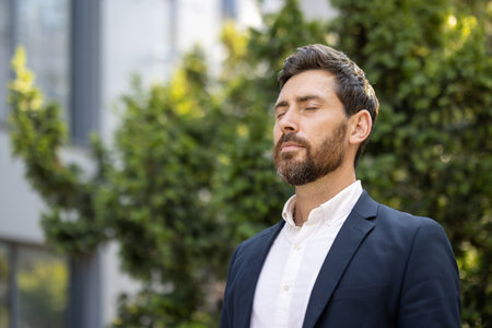 A well-dressed man with eyes closed enjoys a moment of peace outdoors, surrounded by greenery, looking calm and serene.の写真素材