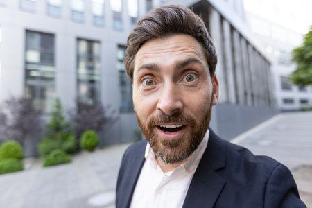A surprised businessman looks directly at the camera outdoors with his mouth open in shock, in front of a modern building.の写真素材