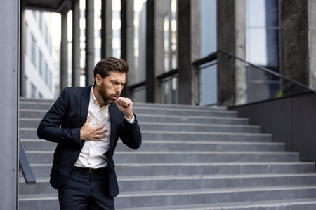 A businessman, clearly unwell, coughs while clutching his chest outdoors. The man is experiencing what seems to be illness or respiratory issues.の写真素材