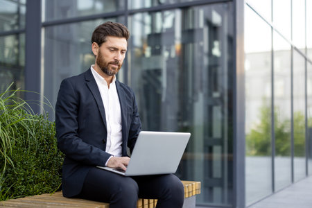 A stylish businessman sits outside a modern building working on his laptop, dressed professionally in a suit.の写真素材