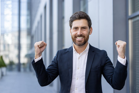 A happy businessman celebrates success with clenched fists outside a modern office building. He is wearing a stylish suit.の写真素材