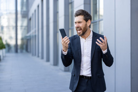 A frustrated businessman gestures while looking at his phone with an angry expression. He appears to be receiving bad news outdoors.の写真素材