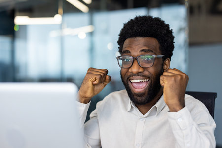 An overjoyed african american man celebrates a win while looking at his laptop screen in an office setting. Success and happiness are reflected on his face.の写真素材