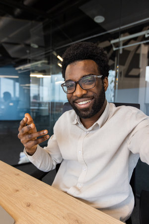 A smiling man in glasses is gesturing, likely in a video call, showcasing enthusiasm in a modern office settingの写真素材