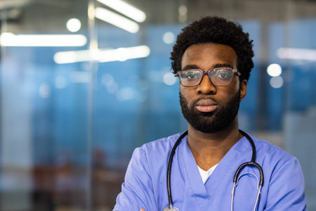 A handsome, young doctor stands confidently in a medical office. His attire includes scrubs and a stethoscope. The office is blurred in the background.の写真素材