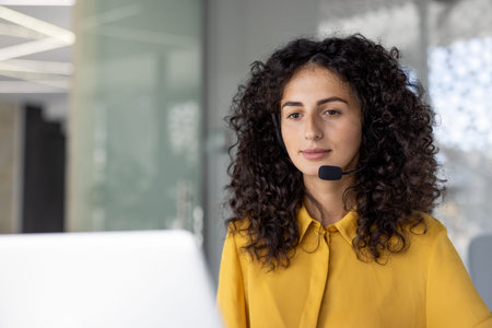 A woman with curly hair wearing a headset, looking attentively at a computer screen, likely...