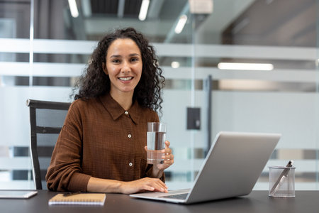 A smiling woman is holding a glass of water and working on a laptop in an office setting.の写真素材